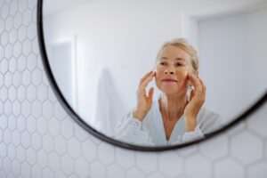 Beautiful senior woman in bathrobe, applying eye patches for puffiness while looking in the mirror